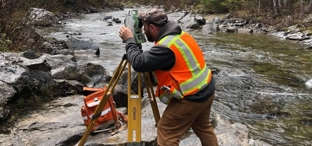 Man in safety vest looking through surveying equipment while standing on flat rocks at the edge of a stream